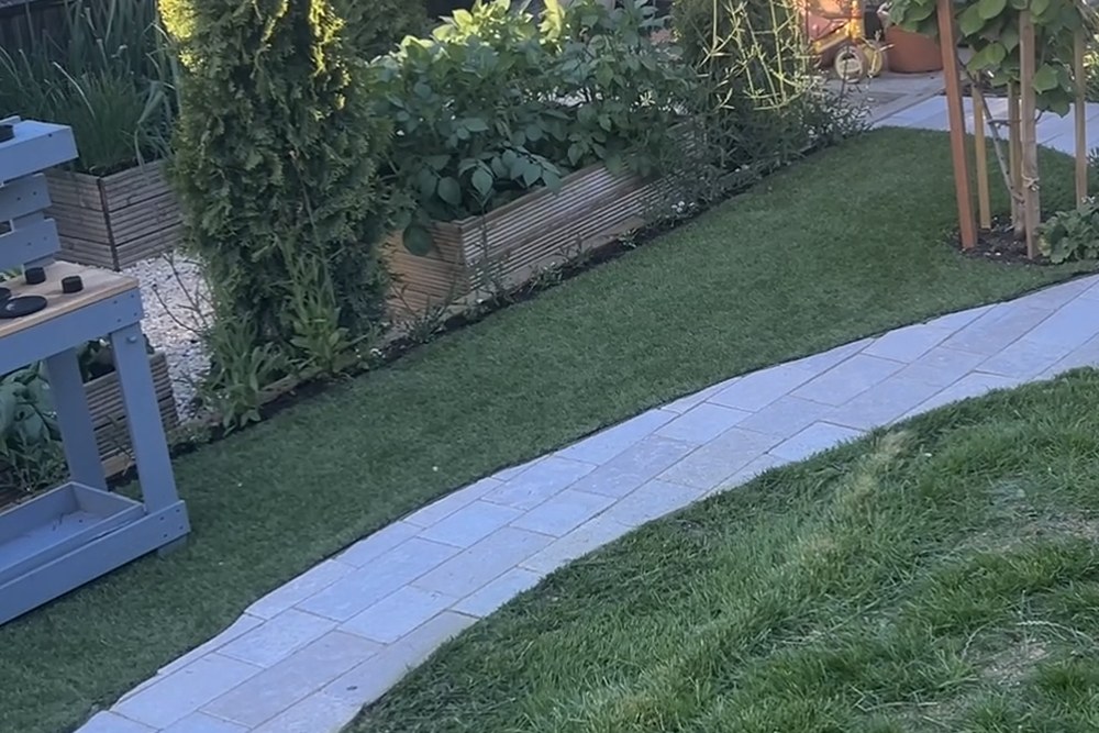 A curved paved path winding through the lawn beside raised beds and a greenhouse